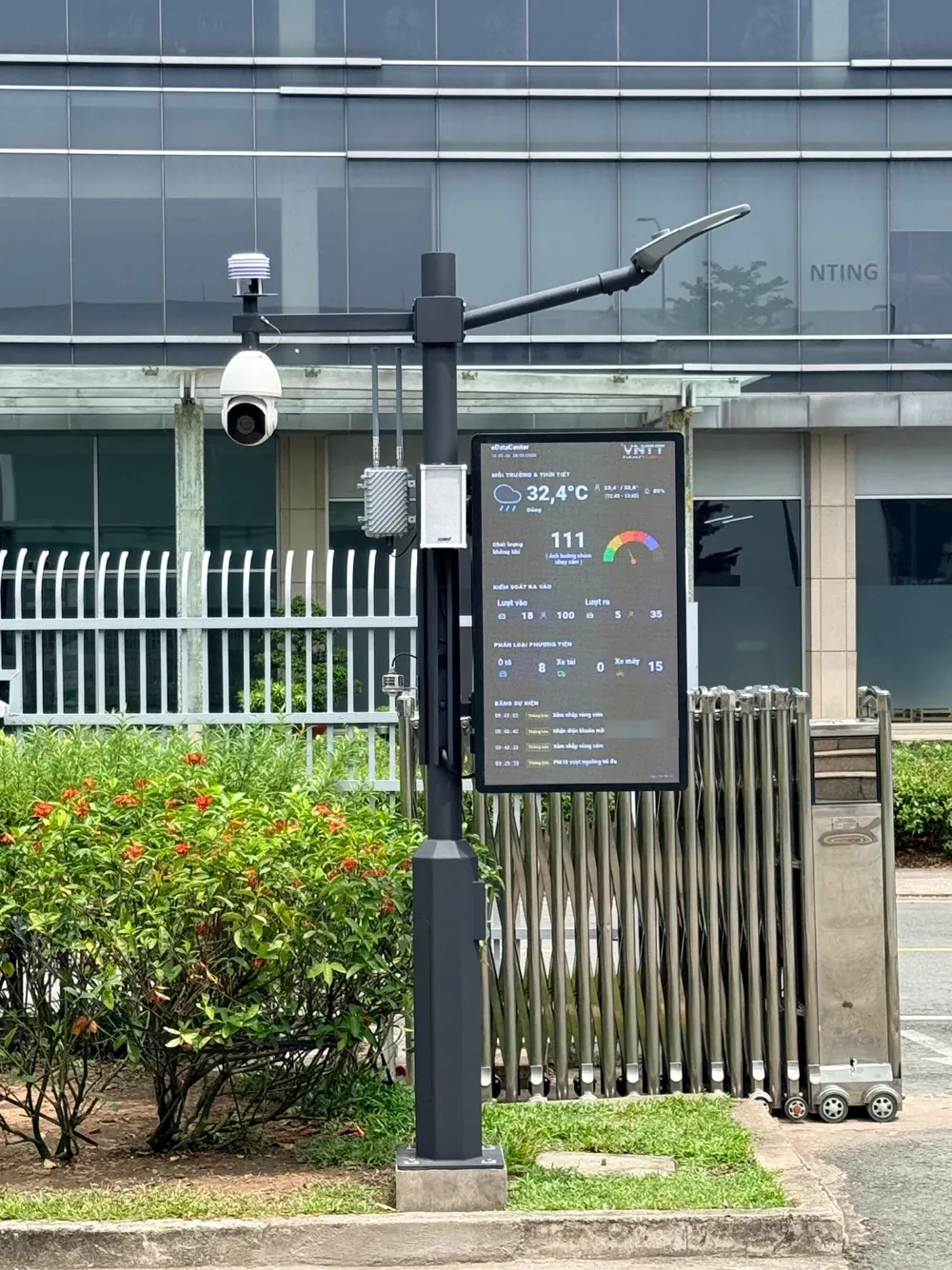 Cận cảnh trụ Smart Pole cạnh cổng xếp: camera, trạm quan trắc, bộ kết nối có anten, loa phát thanh, cảm biến mực nước và màn hình LED đang hiển thị dữ liệu