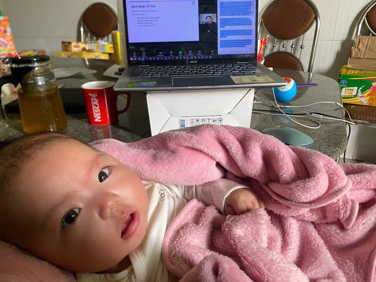 Em bé nằm cạnh máy tính đang mở cuộc họp trực tuyến của mẹ